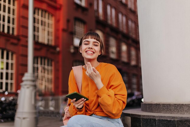 Young shorthaired woman in orange sweatshirt smiles sincerely outside Happy cool girl holds phone and sits outdoors