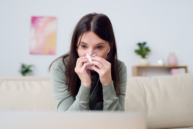 Young sick woman in homewear blowing her nose while sitting on couch in front of laptop and listening to online doctor recommendations