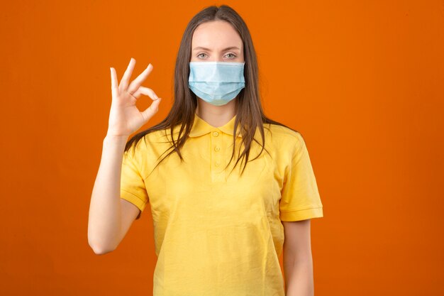 Молодая больная женщина в желтой рубашке поло и медицинской защитной маске, показывая знак ОК, стоя на оранжевом фоне