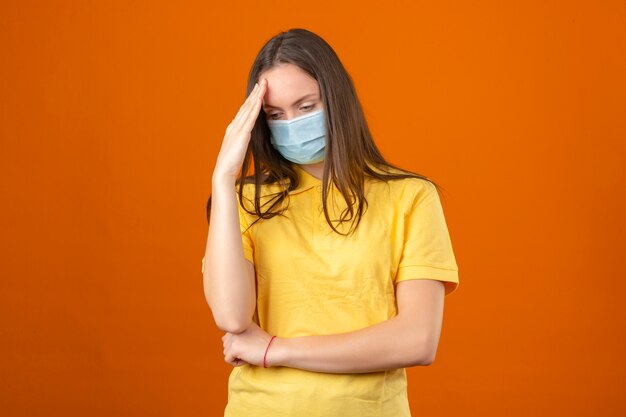 黄色のポロシャツと医療用防護マスクの頭に触れると孤立したオレンジ色の背景に考えて病気の若い女性