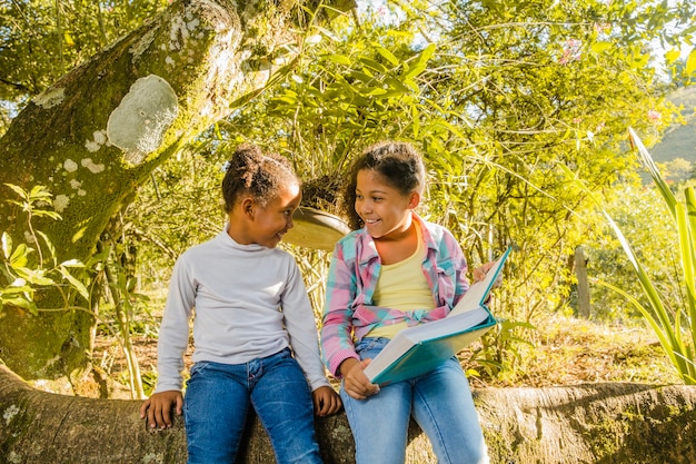 Free photo young sisters with book sitting on a tree