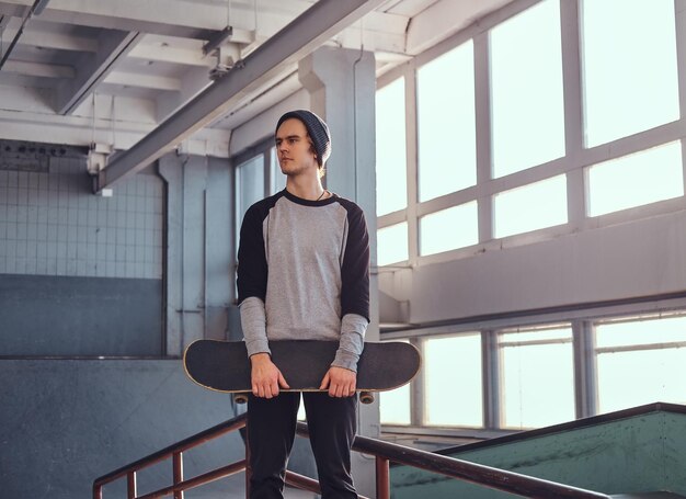 屋内のスケートパークのスケートボードの横に立って、ボードを持って目をそらしている若いスケートボーダー。