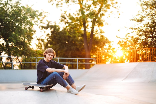 Молодой фигурист в черной футболке и джинсах мечтательно смотрит в камеру, проводя время со скейтбордом в современном скейт-парке на фоне заката
