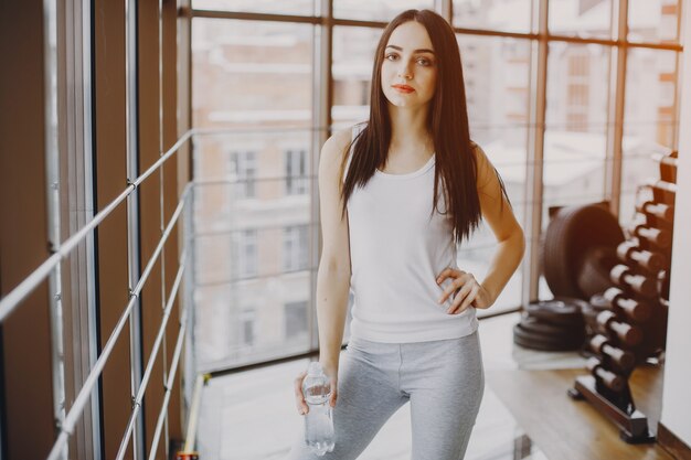 young and skinny girl in a white shirt and gray leggings standing in a gym with bottle 