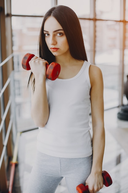 young, skinny girl in a white shirt and gray leggings standing in a gym with red dumbbell