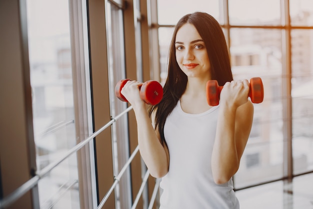 Free photo young, skinny girl in a white shirt and gray leggings standing in a gym with red dumbbell