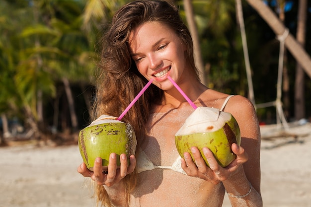 ココナッツを持って、笑顔で、熱帯のビーチで日光浴をしている白いビキニ水着の若い細い女性。