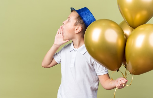 молодой славянский мальчик в синей партийной шляпе держит гелиевые шары и держит руку близко ко рту, зовет кого-то, смотрящего в сторону, изолированную на оливково-зеленой стене с копией пространства