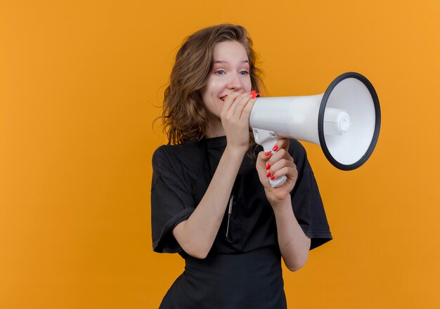 Young slavic female barber wearing uniform looking at side talking by speaker isolated on orange background with copy space