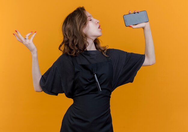 Young slavic female barber wearing uniform singing using mobile phone as microphone keeping hand in air isolated on orange background