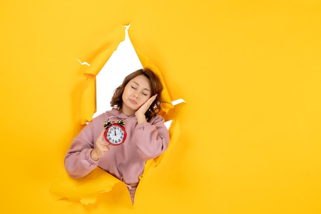 Free photo young sleepy beautiful lady holding clock on yellow torn breakthrought background