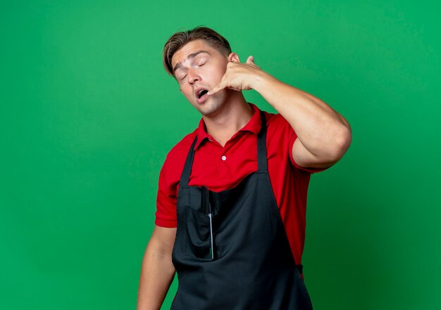 Young sleepy blonde male barber in uniform gestures call hand sign isolated on green space with copy space