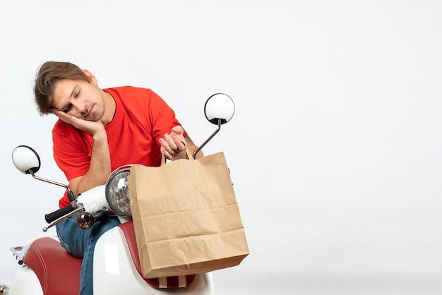 Young sleepy courier man in red uniform sitting on scooter holding paper bag on yellow wall