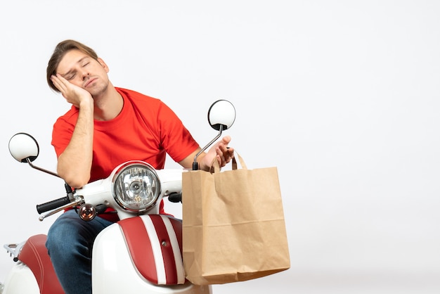 Young sleepy delivery guy in red uniform sitting on scooter holding paper bag on white wall