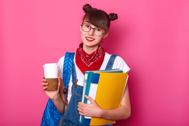 young slender brunette girl in white casual t-shirt, overalls and bandana on neck, holding cup of coffee and folder with documents, posing isolated over rosy. Youngster concept.