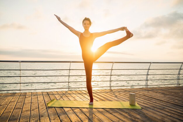 Giovane bella donna attraente sottile che fa yoga al mattino all'alba dal mare, stile di vita sano