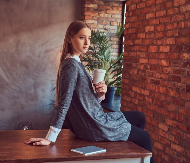 Молодая стройная сексуальная девушка в сером платье держит чашку кофе и сидит