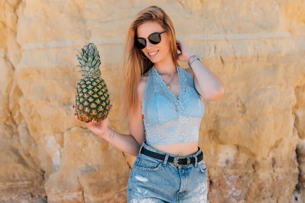 Young slim woman in bikini and sunglasses holding fresh pineapple near the sea