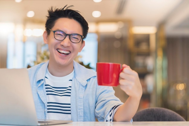 若いスマートなアジア人男性の着用メガネカジュアルシャツは、コーヒーカップの白い歯の笑顔と歓迎の陽気な部屋のインテリアの背景とのこんにちはと手のコミュニケーションを言います