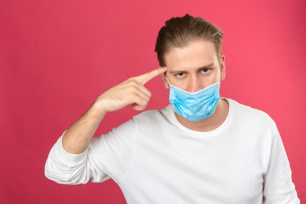 Young smart looking man in medical protective mask pointing on his head over isolated pink background