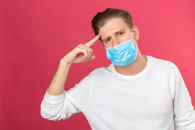 Young smart looking man in medical protective mask pointing on his head over isolated pink background