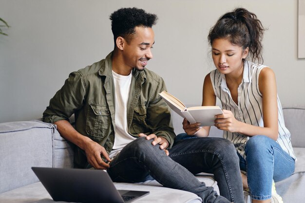 Молодой улыбающийся афроамериканец и симпатичная азиатка мечтательно читают книгу вместе в современном коворкинге