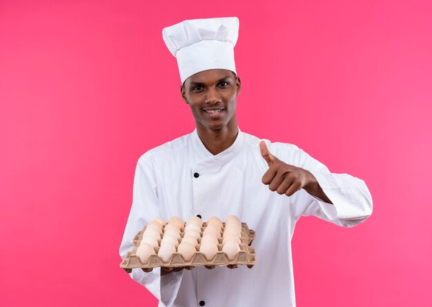 Young smiling afro-american cook in chef uniform holds batch of fresh eggs and thumbs up isolated on pink wall