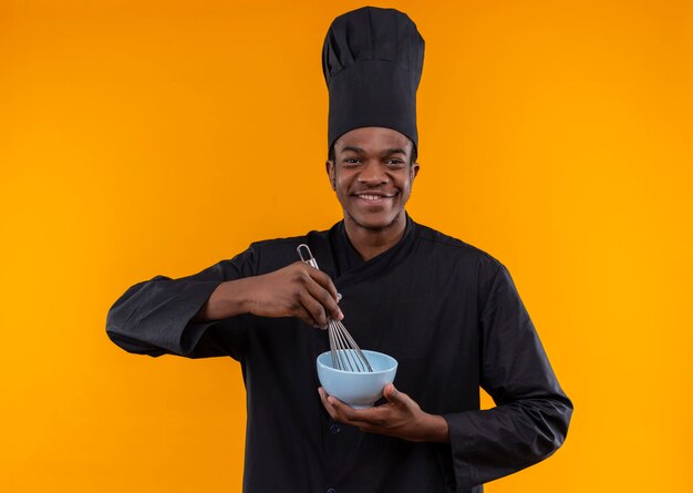 Young smiling afro-american cook in chef uniform holds bowl and whisk isolated on orange wall 