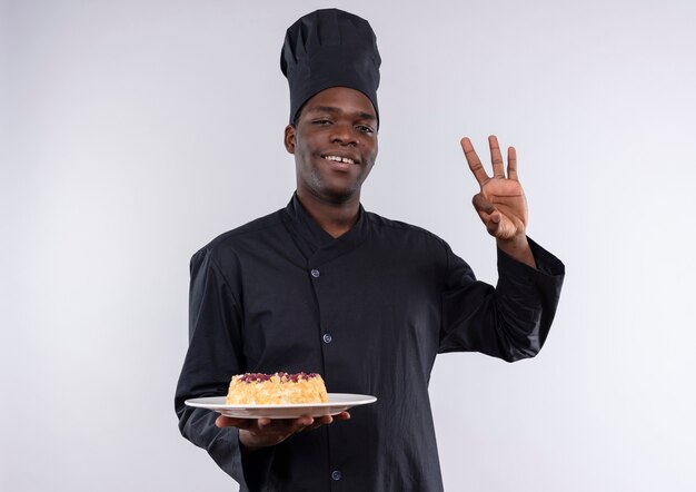 Молодой улыбающийся афро-американский повар в униформе шеф-повара держит торт на тарелке и жестикулирует знак рукой на белом с копией пространства