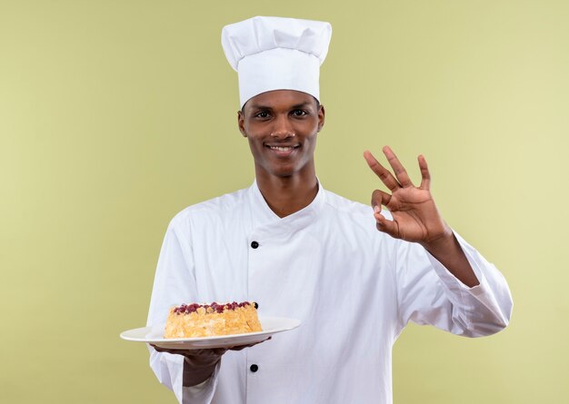 Young smiling afro-american cook in chef uniform holds cake on plate and gwstures ok sign with hand isolated on green wall