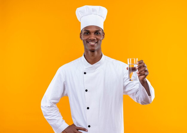 シェフの制服を着た若い笑顔のアフリカ系アメリカ人料理人は、水のガラスを保持し、オレンジ色の壁に隔離された腰に手を置きます