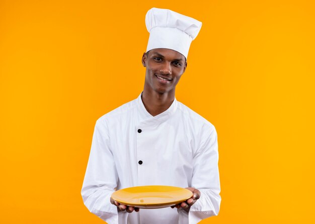 Young smiling afro-american cook in chef uniform holds plate with both hands isolated on orange wall