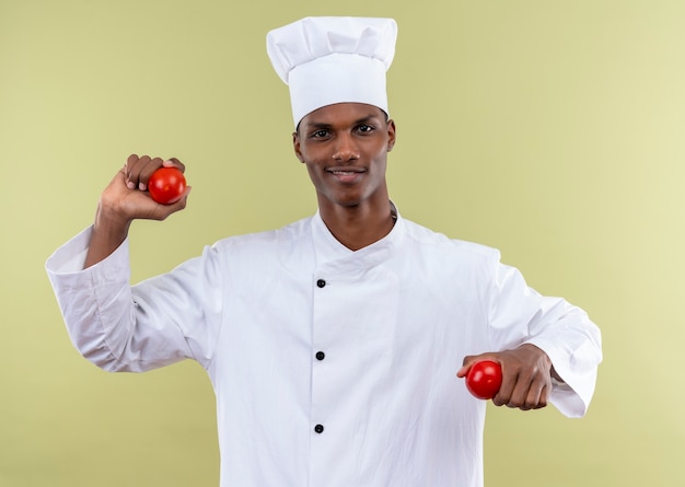 Young smiling afro-american cook in chef uniform holds tomatoes on both hands isolated on green wall