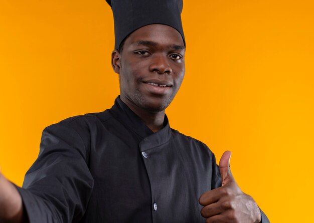 Young smiling afro-american cook in chef uniform pretends to hold camera and thumbs up isolated on orange background with copy space