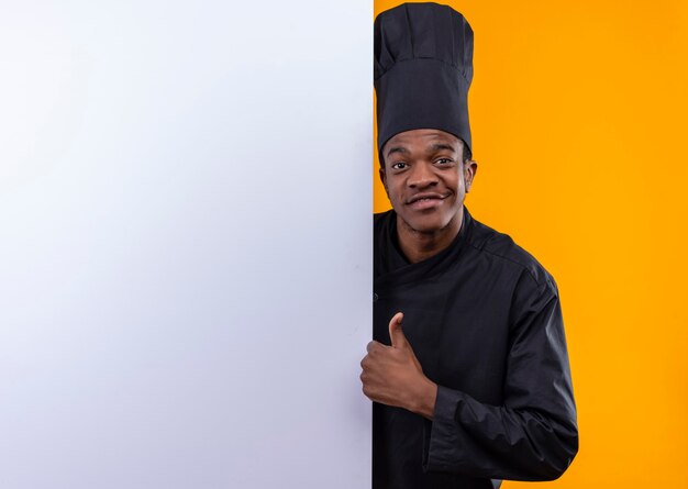 요리사 제복을 입은 젊은 미소 아프리카 계 미국인 요리사는 흰 벽 뒤에 서서 오렌지 벽에 고립 된 엄지 손가락