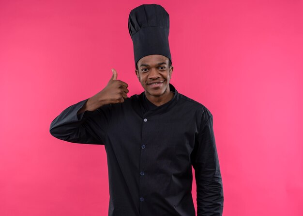 Молодой улыбающийся афро-американский повар в униформе шеф-повара показывает палец вверх на розовой стене