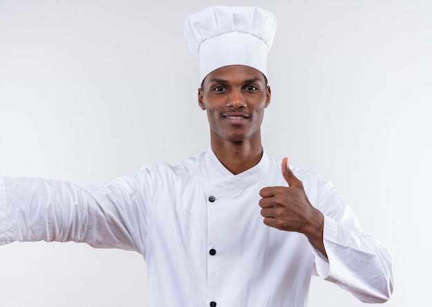 Young smiling afro-american cook in chef uniform thumbs up isolated on white wall