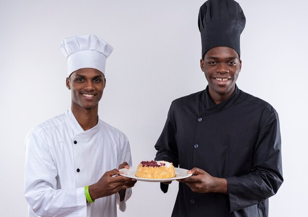 Молодые улыбающиеся афро-американские повара в униформе шеф-повара держат торт на тарелке вместе, изолированные на белой стене