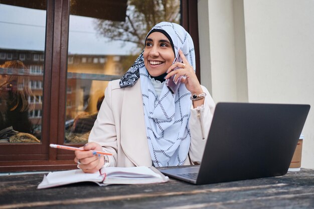 Young smiling Arabic woman in hijab happily talking on smartphone while working on laptop on street