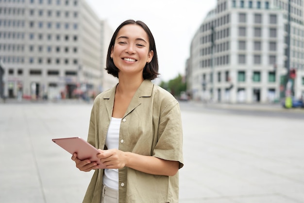 無料写真 日光の下で通りに立っているタブレットを持つ若い笑顔のアジア女性