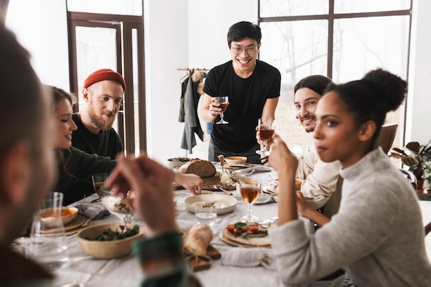 Giovane uomo asiatico sorridente in occhiali e maglietta nera che tiene felicemente un bicchiere di vino in mano gruppo di amici internazionali positivi attraenti che trascorrono del tempo insieme a pranzo nell'accogliente caffè