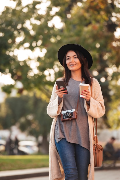 Молодая улыбающаяся азиатская женщина в стильной одежде, держащей смартфон и кофе, чтобы пойти на открытом воздухе