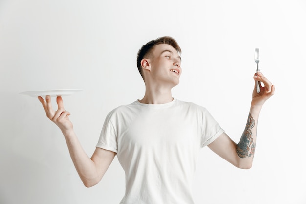 Young smiling attractive caucasian guy holding empty dish and fork isolated on grey background. Copy space and mock up. Blank template background.