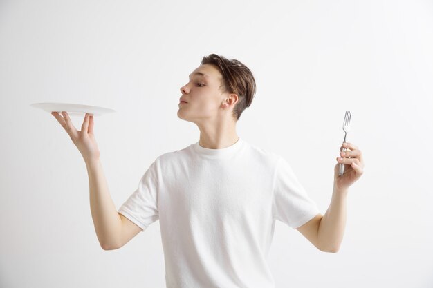 Young smiling attractive caucasian guy holding empty dish and fork isolated on grey background. Copy space and mock up. Blank template background.