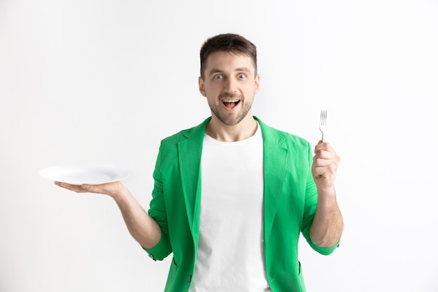 Young smiling attractive caucasian guy holding empty dish and fork isolated on grey background. Copy space and mock up. Blank template background.