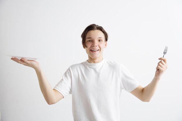 Young smiling attractive caucasian guy holding empty dish and fork isolated on grey background. Copy space and mock up. Blank template background.