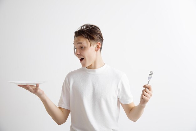 Young smiling attractive guy holding empty dish and fork isolated on grey wall.