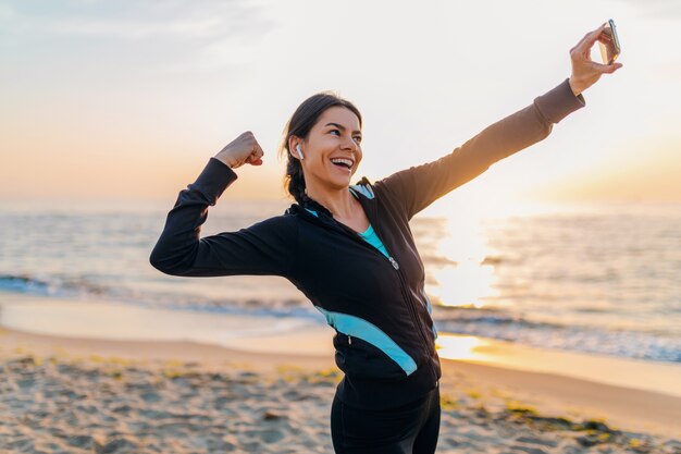 Young smiling attractive slim woman doing sport exercises on morning sunrise beach in sports wear, healthy lifestyle, listening to music on earphones, making selfie photo on phone looking strong