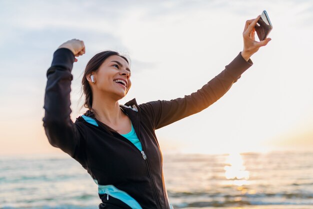 Молодая улыбающаяся привлекательная стройная женщина делает спортивные упражнения на утреннем пляже восхода солнца в спортивной одежде, здоровом образе жизни, слушает музыку в наушниках, делает селфи-фото на телефоне, выглядит сильным