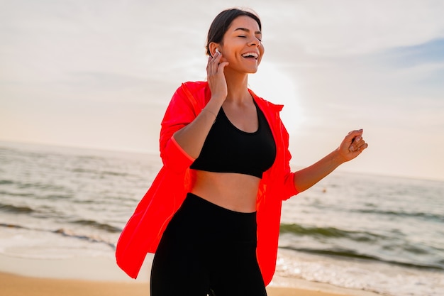 Young smiling attractive slim woman doing sports in morning sunrise dancing on sea beach in sports wear, healthy lifestyle, listening to music on earphones, wearing pink windbreaker jacket, having fun